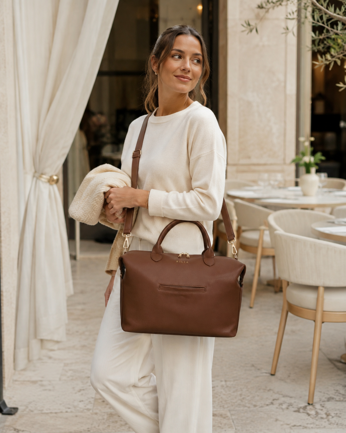 Frau trägt die RILIA Signature Bag in Cognac mit Crossbody-Gurt vor einem mediterranen Restaurant